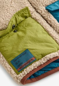 Chaqueta de forro polar beige con paneles de tela verdes y azules. Presenta una etiqueta que dice "Mañana es una nueva aventura." Texturizada y cálida.