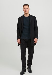 Svart yllefrack med skräddarsydd passform, bärs över en texturerad mörkblå tröja och svarta kordbyxor. Svarta snörboots fulländar looken.