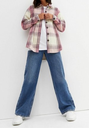 Persona con vaqueros anchos azules, zapatillas blancas, camisa blanca y chaqueta de cuadros rosa con botones, de pie frente a un fondo blanco.