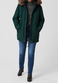 Parka verte à capuche avec bordure en fourrure, dotée d'une fermeture éclair, d'une taille à cordon et de poches latérales, associée à un jean bleu et des bottes noires.