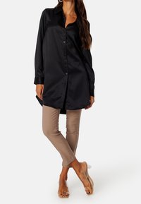 Camisa negra de satén de manga larga con botones, combinada con pantalones beige de corte slim y sandalias transparentes, presentando una textura suave y un ajuste a medida.