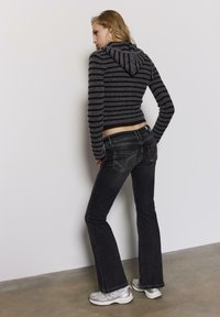 Trenirka z dolgimi rokavi s kapuco v sivih in rjavih črtah, skrajšana, kombinirana s temnimi zvončastimi džins hlačami z dekorativnim vezenjem na zadnjih žepih.