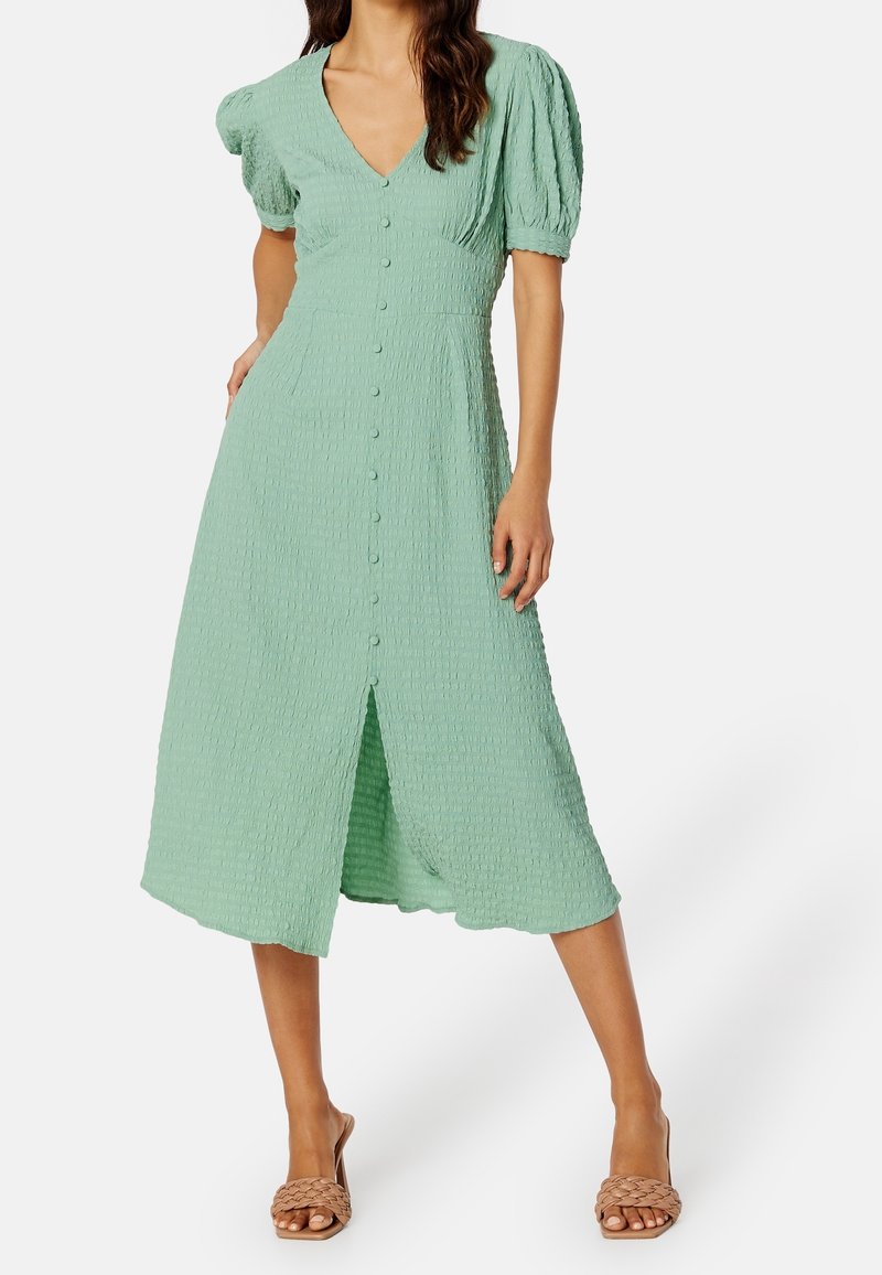 Femme portant une robe midi texturée vert clair avec des manches bouffantes, des boutons sur le devant et une fente avant, associée à des sandales tissées couleur beige.