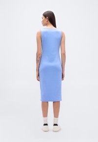 Ανοιχτό μπλε φόρεμα χωρίς μανίκια με εφαρμοστή σιλουέτα, που φτάνει μέχρι τη μέση της γάμπας, κατασκευασμένο από απαλό ύφασμα. Φοριέται με λευκές κάλτσες και αθλητικά παπούτσια.