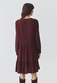Robe imprimée bordeaux à manches longues, avec des volants en cascade à l'ourlet et un col rond, présentant un motif géométrique en bleu.