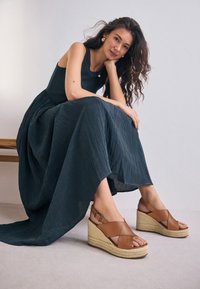 Sandales compensées en cuir marron avec un design à brides croisées, une semelle intermédiaire en jute tressé et une finition lisse et brillante. Le modèle porte une robe sombre fluide.