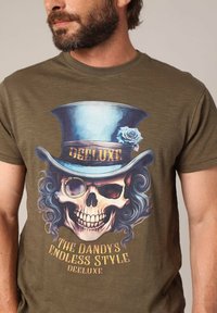 T-shirt vert olive avec un graphique d'un crâne portant un haut-de-forme bleu orné d'une rose, avec le texte "Le style infini du dandy" en dessous.