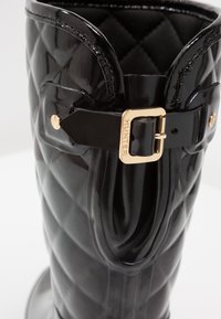 Gros plan d'une botte en caoutchouc matelassée noire avec une sangle à boucle dorée étiquetée "HUNTER" près du bord supérieur.