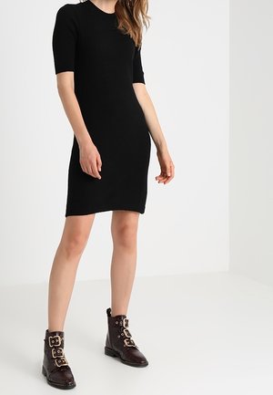Zwarte gebreide jurk met korte mouwen, aansluitende pasvorm en ronde halslijn, gecombineerd met bruine enkellaarsjes met gouden gespen.