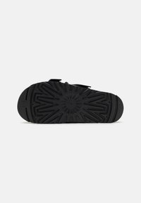 Sandale noire avec une semelle en caoutchouc texturée pr�ésentant un motif en rayons de soleil, conçue pour l'adhérence et la stabilité. Des sangles ajustables complètent le design.