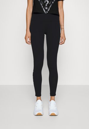 Black leggings made of stretchy material, featuring a high waist and a logo accent on the left leg, paired with white sneakers.