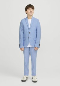 Costume bleu clair avec un bouton unique, revers crantés et poches avant, associé à un t-shirt blanc et des baskets noir et blanc.