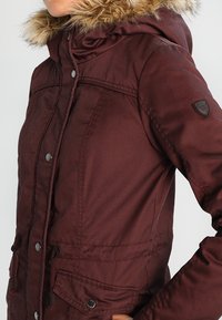 Bordowa kurtka z kapturem wykończonym sztucznym futrem, z zamkiem błyskawicznym i zatrzaskiem, dwoma przednimi kieszeniami oraz małą naszywką z logo na rękawie.