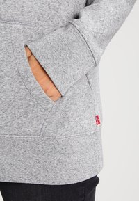 Sudadera gris con un material de forro polar texturizado, que cuenta con un bolsillo lateral, puños de canalé y una pequeña etiqueta de logo roja en el dobladillo inferior.