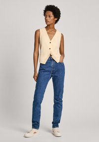 Κρεμ αμάνικο γιλέκο με βαθύ V-λαιμόκοψη, διαθέτει τρία καφέ κουμπιά. Συνδυάζεται με ψηλόμεσα μπλε τζιν και μπεζ υποδημάτων.