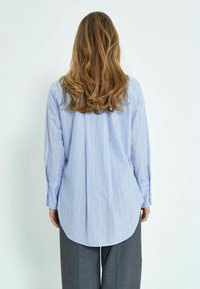 Camicia a maniche lunghe a righe blu con orlo curvo, caratterizzata da righe verticali bianche e arancioni e polsini con bottoni. Indossata con pantaloni grigi.