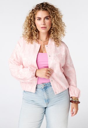 Vrouw met krullend blond haar die een roze textuurjasje, roze top, lichtblauwe spijkerbroek, gouden ketting, ringen en armbanden draagt tegen een witte achtergrond.