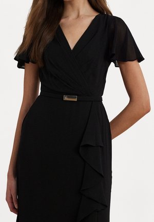 Frau trägt ein schwarzes Wickelkleid mit Schmetterlingsärmeln, einem V-Ausschnitt, einem dünnen Gürtel mit kleiner goldener Schnalle und einem Rüschendetail am Rock.