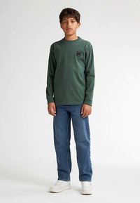 Camiseta de algodón verde de manga larga con cuello redondo y pequeño parche con el logo; combinada con vaqueros azules y zapatillas blancas.