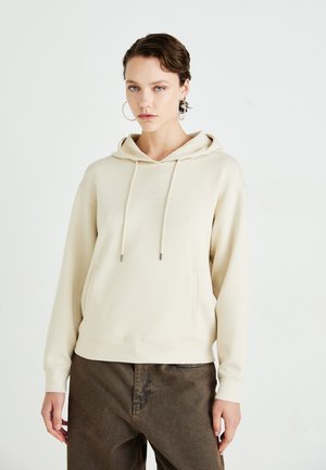 Crème hoodie met een aantrekkoord, een voorzak en een reliëflogo. Draagt bij een paar bruine gestructureerde broek. Gladde stof en een ontspannen pasvorm.