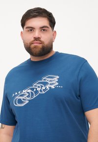 Modrá bavlněná tričko s bílým grafickým potiskem s textem "JACK & JONES 1990" a abstraktními designovými prvky. Dobrý střih, kulatý výstřih.