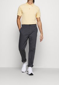 Man wearing a pale yellow short-sleeve polo shirt, dark gray rolled-up pants, white socks, and white athletic shoes walking.