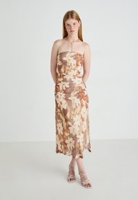 Mouwloze, bloemenmidi-jurk in bruin en crème, met geplooide textuur en een getailleerd silhouet. Gecombineerd met sandaaltjes met bandjes.