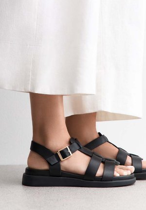 Des sandales en cuir noir présentent des larges lanières, un accent de boucle et une semelle plate. Le design est ouvert sur l'avant, laissant visible la peau des pieds.