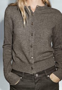 Cardigan grigio lavorato a maglia con scollatura rotonda, cinque bottoni metallici sul davanti e polsini a coste. Indossato sopra pantaloni scuri.