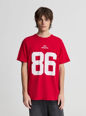 Młody mężczyzna w czerwonej koszulce z napisem „NYC ATHLETIC” i dużym białym numerem 86 stoi na jasnym, jednolitym tle.