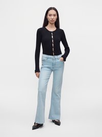 Femme portant un haut noir à manches longues boutonné et un jean flare bleu clair taille haute, debout avec une main dans la poche.