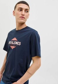 Tamsiai mėlynos spalvos medvilninė marškinėliai su trumpomis rankovėmis, su centriniu logotipo grafiku raudona ir balta spalvų su vintage dizainu.