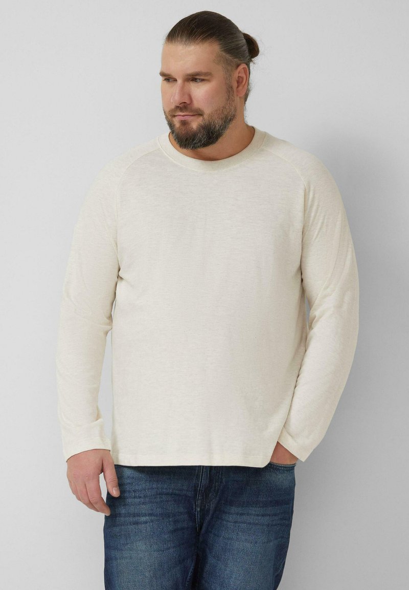 Jasnokremowa koszulka z długim rękawem, okrągłym wycięciem na szyję, luźnym krojem i teksturowaną tkaniną, zestawiona z ciemnoniebieskimi dżinsami.