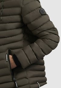 Kurdinātā jakas tumši olīvzaļā krāsā, ar siltinātu dizainu, horizontālu šuvju rakstu, rāvējslēdzēja kabatu un melnu logo uzplecu. 