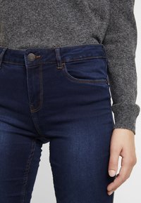 Mörkblåjeans med en slät textur, med en klassisk design, kontrasterande sömmar och en metallknappstängning i midjan.