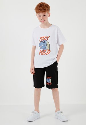 Punapäine poiss, kes kannab valgeid tossusid, valget T-särki ja musti lühikesi pükse sinise dinosaurusemotiivi ja tekstiga „Stay Wild“, seisab valgel taustal.