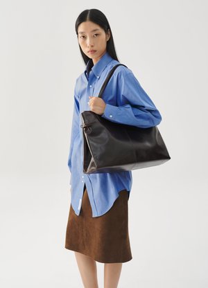 Brown leather tote bag with a smooth texture, secured by a zip, held by a person wearing a blue button-up shirt and brown suede skirt.