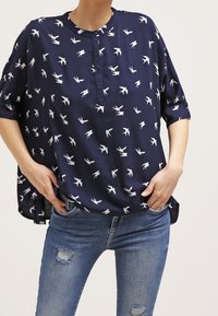 Blouse bleu marine avec motifs d'oiseaux blancs, manches courtes et col rond. Tissu doux et léger avec une coupe décontractée.