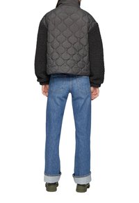 Persoon in een zwarte gewatteerde jas met textuur op de mouwen, blauwe opgerolde spijkerbroek en olijfgroene laarzen, staand met de rug naar de kijker.