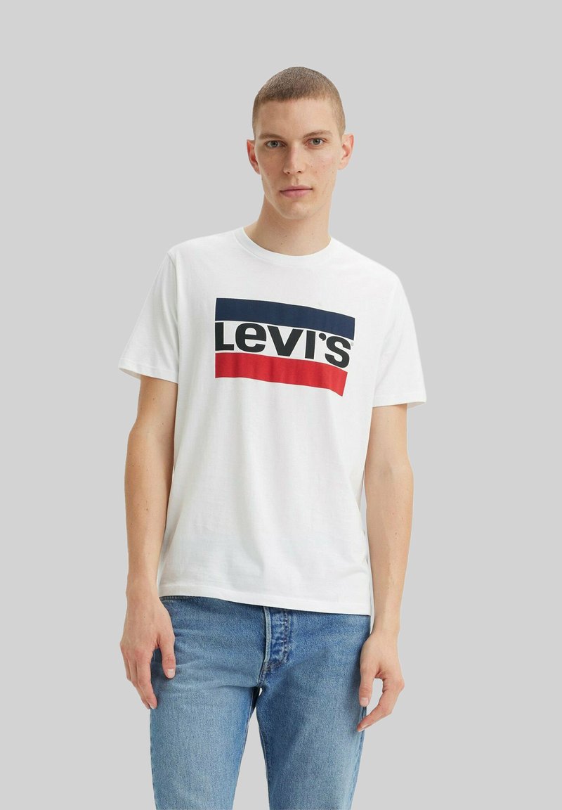 Jonge man in een witte Levi's t-shirt met marineblauwe en rode horizontale strepen en blauwe jeans, staand tegen een effen grijze achtergrond.