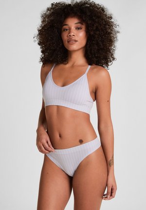 Vrouw met krullend haar die een lichtgrijze gestreepte beha en bijpassend ondergoed poseert tegen een effen grijze achtergrond.