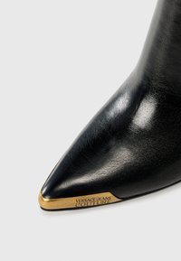 Sorte lærpointe støvler med en glansfuld finish, med et guldfarvet metalaccent på hælen indgraveret med "VERSACE JEANS COUTURE."