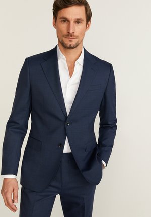 Marineblauw pak van gestructureerde stof, voorzien van een twee-knopen sluiting, notch revers en twee voorkeurszakken, gecombineerd met een witte shirt.