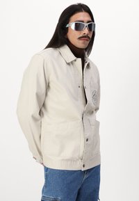 Beige, button-up jacket with a collar, two front pockets, and a chest logo. Worn over a brown turtleneck; paired with blue denim.