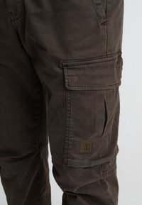 Cargo pants in donkerbruin katoen met patchzakken, zichtbare stiksels en een vierkante logopatch op de linkerdij. Getextureerde stof.