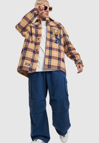 Plaid flannel shirt in yellow, blue, and red with button closure and logo detail; paired with navy cargo pants; set against a neutral background.