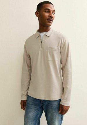 Polo a maniche lunghe beige con colletto a mezza zip, taschino singolo sul petto e una morbida texture di cotone. Indossato con jeans blu.