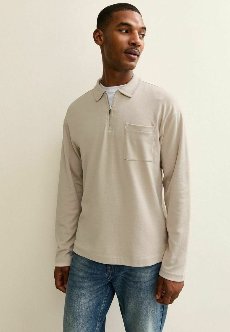 Camisa polo de manga larga beige con cuello de media cremallera, bolsillo en el pecho y una textura suave de algodón. Combinada con jeans azules.