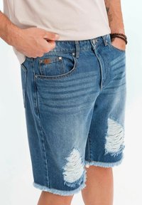 Blå denimshorts med slitna hål och fransade fållar. Har fem fickor och en läderlapp på midjebandet.