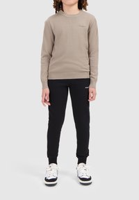 Beige gebreide trui met geribbelde ronde hals, gecombineerd met zwarte sweatpants met een logo. Model draagt witte en zwarte sneakers.